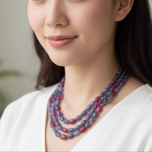 May include: A three-strand necklace with oval beads in alternating blue and red hues. The necklace is worn over a white top, creating a contrast. The beads are polished and have a subtle shine, reflecting light. A piece of jewellery.