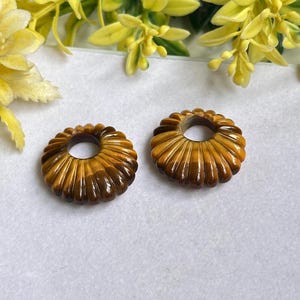 May include: Two circular, fluted beads crafted from brown and gold tiger's eye gemstone. Each bead features a smooth, polished surface with a distinct chatoyant luster and a central hole, showcasing the natural banded patterns of the stone.