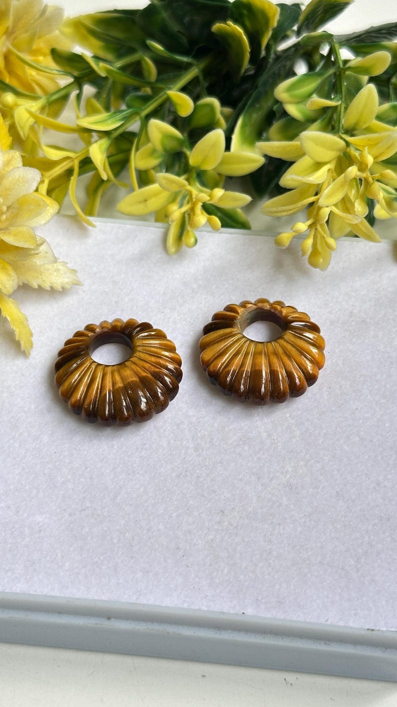 May include: Two circular, fluted beads crafted from brown and gold tiger's eye gemstone. Each bead features a smooth, polished surface with a distinct chatoyant luster and a central hole, showcasing the natural banded patterns of the stone.