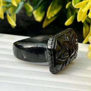 May include: A black ring with a rectangular face featuring a carved floral design. The ring has a smooth, polished band and a detailed, textured face. The ring is set against a background of yellow flowers and green foliage.