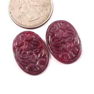 Puede incluir: Dos piedras preciosas ovaladas de rubí tallado con diseños florales. Las piedras preciosas son de color rojo intenso y están junto a una moneda de veinticinco centavos para comparar el tamaño. Sobre un fondo blanco.