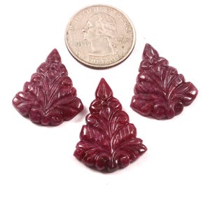 May include: Three carved ruby gemstones in a leaf design, with a quarter coin for size comparison. The gemstones are a deep red color and have intricate details. The gemstones are on a white background.