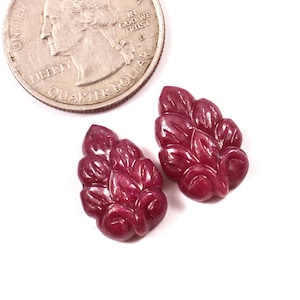 Puede incluir: Dos gemas de rubí talladas con un diseño en forma de hoja, junto a una moneda de veinticinco centavos. Las gemas son de color rojo intenso y tienen un acabado pulido. La moneda es plateada y tiene las palabras "Quarter Dollar".