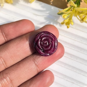 May include: A carved, circular, deep red gemstone in the shape of a rose. The gemstone is held in a hand, with the rose design clearly visible. The petals are intricately carved, and the stone has a polished finish. Yellow flowers are in the background.