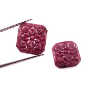 Peut inclure: Deux pierres précieuses en rubis sculptées. L'une est tenue par une pince, l'autre repose sur une surface blanche. Chaque pierre est de forme rectangulaire avec un motif floral sculpté. Les rubis sont d'un rouge profond et riche.