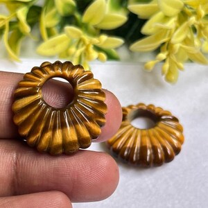 May include: Two circular tiger's eye gemstone beads featuring a carved, fluted, or ribbed radial pattern. Each bead has a smooth, polished finish with a distinct golden-brown chatoyant luster and a central circular hole for threading.