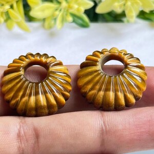 May include: Two circular beads carved from golden-brown tiger's eye gemstone. Each bead features a radial, fluted, or ribbed pattern radiating from a central circular hole, showcasing the natural chatoyant luster and banded texture of the stone.