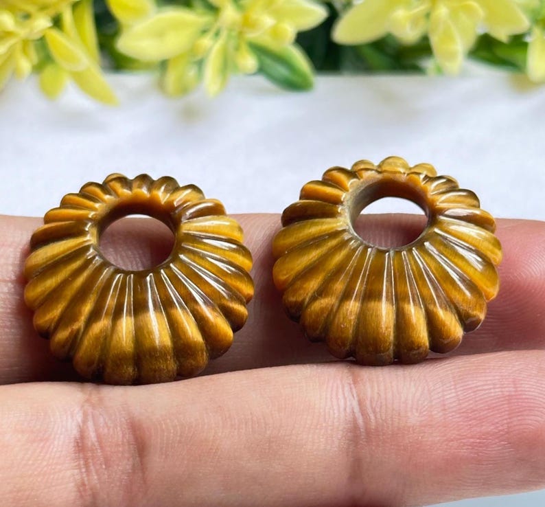 May include: Two circular beads carved from golden-brown tiger's eye gemstone. Each bead features a radial, fluted, or ribbed pattern radiating from a central circular hole, showcasing the natural chatoyant luster and banded texture of the stone.