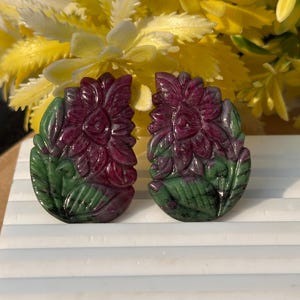May include: Two carved gemstone pendants. Each pendant features a flower design with deep red petals and green leaves. The gemstones are set against a background of yellow and white flowers.