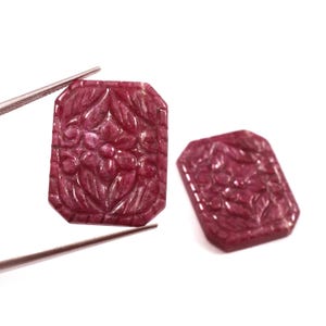 May include: Two carved ruby gemstones with floral designs. The rectangular stones are a deep red colour and feature detailed carvings of flowers and leaves. One stone is held by tweezers.