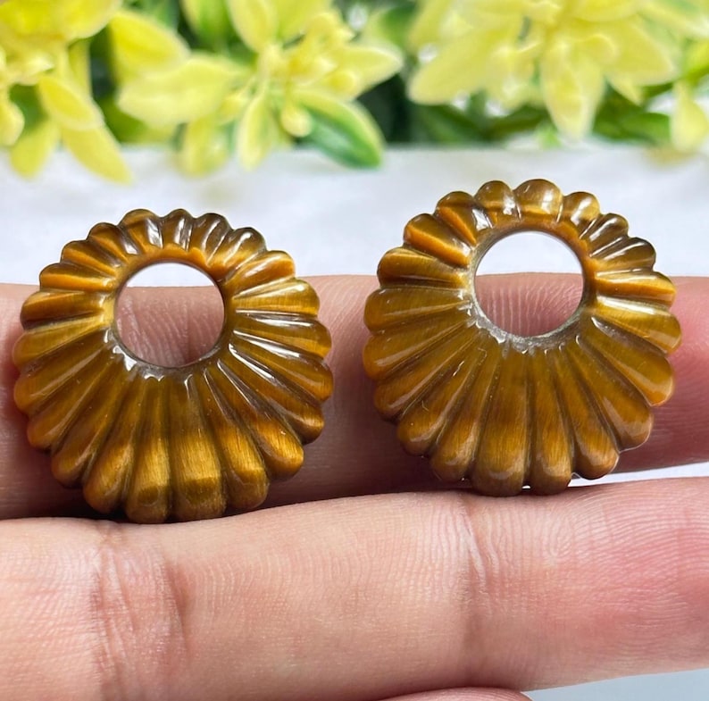 May include: Two circular tiger's eye gemstone components featuring a carved, fluted, or sunburst-like radial pattern. Each piece has a smooth, polished finish with a distinct chatoyant golden-brown luster and a centered circular cutout.