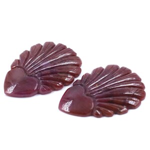 Pode incluir: Duas pedras preciosas em forma de coração, esculpidas em um tom bordô profundo. Cada gema apresenta uma base em forma de coração com um design em forma de leque. As gemas estão em um fundo branco.