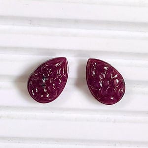 Puede incluir: Dos cabujones en forma de lágrima de un tono rojo magenta intenso. Cada pieza presenta un patrón floral tallado detallado en su superficie, mostrando pétalos y hojas intrincados grabados en el material de piedra preciosa pulida.