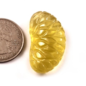May include: A carved, yellow gemstone shaped like a citrus fruit segment. The gemstone is next to a U.S. quarter for size comparison. The gemstone has detailed leaf-like carvings across its surface, and is set against a white background.