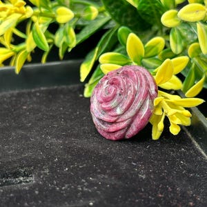 Puede incluir: Una gema tallada en forma de rosa, en tonos rosa y blanco, descansa sobre una superficie oscura, con follaje amarillo y verde en el fondo. El intrincado diseño y las variaciones de color de la gema crean una pieza visualmente atractiva.