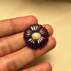 May include: A deep purple, carved amethyst gemstone shaped like a flower with rounded petals. The center of the floral carving features a smooth, oval-shaped, iridescent white opal cabochon that displays subtle flashes of blue and orange light.