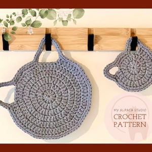Op de afbeelding: Twee grijze gehaakte ovenwanten met handvatten die aan een houten rek hangen. De grotere ovenwant heeft een lusvormig handvat, terwijl de kleinere een enkel handvat heeft. De tekst "My Alpaca Studio Crochet Pattern" is zichtbaar onder de ovenwanten.