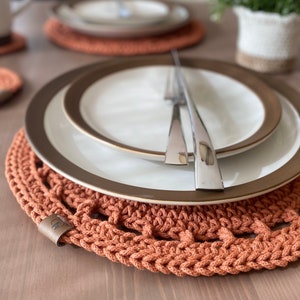 May include: A table setting featuring a crocheted orange placemat with a matching coaster. White plates with a gold rim and silverware are placed on the placemat. The setting is on a light brown wooden table.