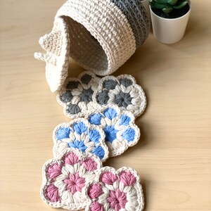 May include: Set of crocheted coasters in various colors, including gray, blue, and pink. The coasters are made with a flower-shaped design and are arranged on a wooden surface. A crocheted basket with a lid is also visible in the background.