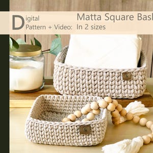 May include: Two beige crocheted square baskets of different sizes, one with a white cloth inside. A wooden bead garland and a candle in a glass jar are also present. The text reads "Digital Pattern + Video: Matta Square Basket In 2 sizes."