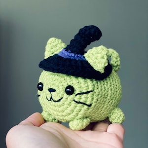 Puede incluir: Un gato de crochet verde con un sombrero de bruja negro con un borde morado. El gato tiene ojos negros y una sonrisa cosida.