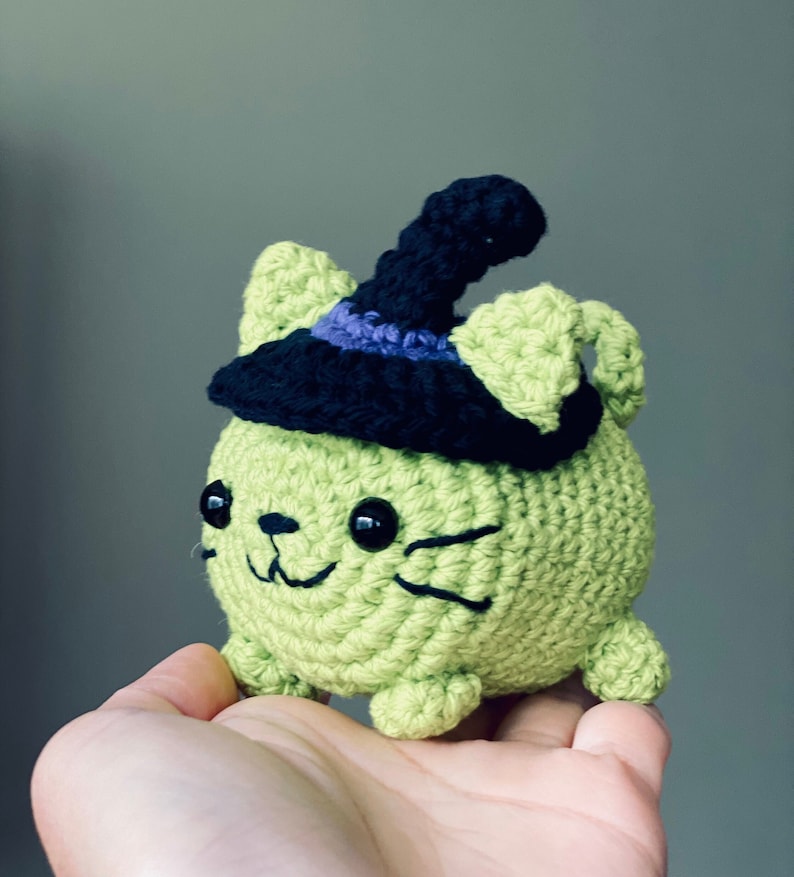 Puede incluir: Un gato de crochet verde con un sombrero de bruja negro con un borde morado. El gato tiene ojos negros y una sonrisa cosida.