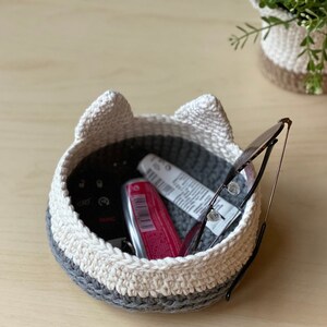 May include: A crocheted cat-shaped basket with white and gray yarn. The basket is filled with various items, including a pair of glasses, a tube of lip balm, and a key.