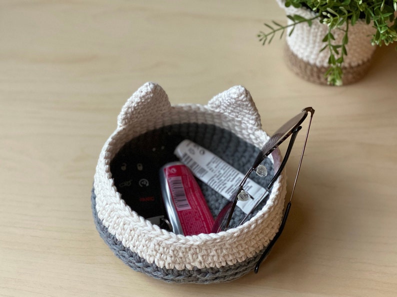 May include: A crocheted cat-shaped basket with white and gray yarn. The basket is filled with various items, including a pair of glasses, a tube of lip balm, and a key.