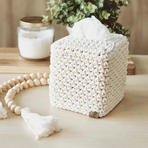 May include: A square, off-white macrame tissue box cover. The cover has a woven pattern and a small, brown tag on the bottom. White tissues are visible at the top. A wooden bead garland with tassels and a candle are in the background.