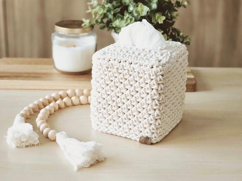 May include: A square, off-white macrame tissue box cover. The cover has a woven pattern and a small, brown tag on the bottom. White tissues are visible at the top. A wooden bead garland with tassels and a candle are in the background.