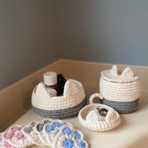 May include: A set of crocheted bathroom accessories in white and gray. The set includes a small round container with a lid, a larger round container with a lid, a small round dish, and six small heart-shaped pads. The pads are in shades of pink and blue.