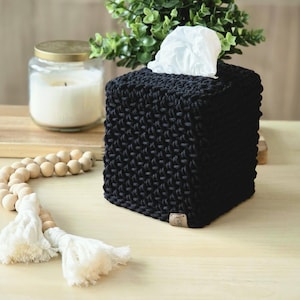 May include: A black, square tissue box cover with a textured knit design. White tissues are visible at the top. A wooden beaded garland with white tassels and a candle in a glass jar are also in the image.