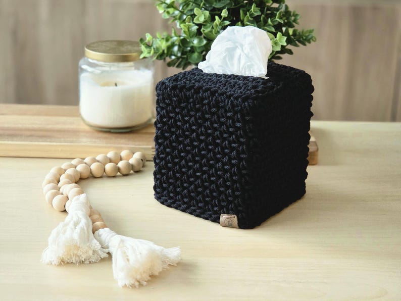 May include: A black, square tissue box cover with a textured knit design. White tissues are visible at the top. A wooden beaded garland with white tassels and a candle in a glass jar are also in the image.