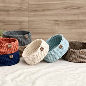 Op de afbeelding: Vijf gehaakte manden in verschillende kleuren, waaronder grijs, oranje, donkerblauw, crème en lichtblauw. De manden zijn allemaal rond en hebben een klein bruin leren label aan de zijkant.