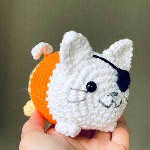 Puede incluir: Un gato de crochet blanco con un cuerpo a rayas naranja y amarillo. El gato tiene un parche en el ojo negro y una sonrisa cosida.