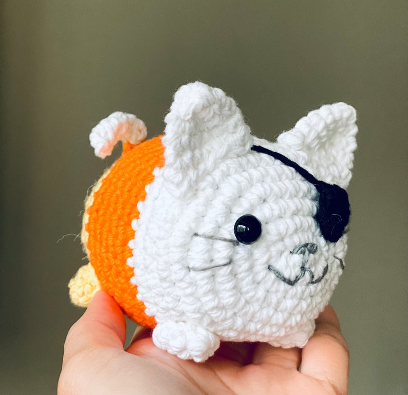 Puede incluir: Un gato de crochet blanco con un cuerpo a rayas naranja y amarillo. El gato tiene un parche en el ojo negro y una sonrisa cosida.