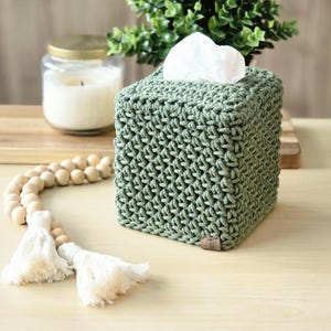 May include: A sage green, square tissue box cover made of a knitted material. White tissues are visible at the top. A wooden bead garland with white tassels and a lit candle in a glass jar are also in the image.
