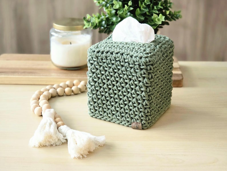 May include: A sage green, square tissue box cover made of a knitted material. White tissues are visible at the top. A wooden bead garland with white tassels and a lit candle in a glass jar are also in the image.