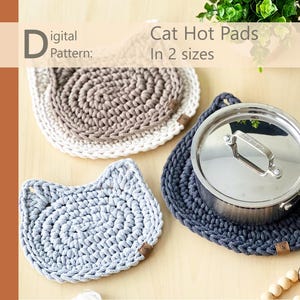 May include: Two crocheted cat-shaped hot pads in gray and beige. The larger pad is on a wooden surface with a silver pot on top of a smaller, dark gray crocheted cat-shaped hot pad. The text "Cat Hot Pads In 2 sizes" is above the image.