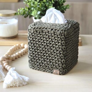 May include: A square, olive green tissue box cover with a textured, woven design. White tissues peek out from the top. A glass jar candle with a gold lid, a wooden bead garland, and greenery are in the background.
