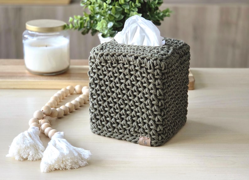 May include: A square, olive green tissue box cover with a textured, woven design. White tissues peek out from the top. A glass jar candle with a gold lid, a wooden bead garland, and greenery are in the background.