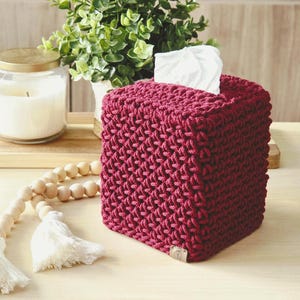 May include: A burgundy, square tissue box cover with a textured, woven design. White tissues peek out from the top opening. A lit candle in a glass jar and a decorative wooden bead garland with tassels are in the background.
