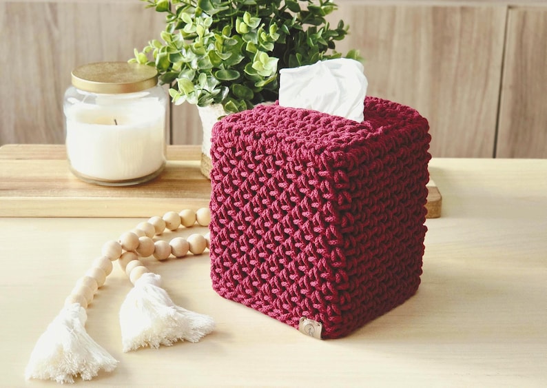 May include: A burgundy, square tissue box cover with a textured, woven design. White tissues peek out from the top opening. A lit candle in a glass jar and a decorative wooden bead garland with tassels are in the background.