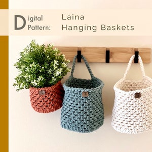 May include: Three hanging baskets made of macrame cord, in beige, teal, and terracotta, are hanging from a wooden rack with black hooks. The baskets are empty, but the terracotta basket has a small potted plant inside.