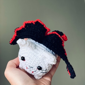 Puede incluir: Un gato blanco de crochet con una capa de crochet negra y roja. El gato tiene ojos negros y una boca roja.