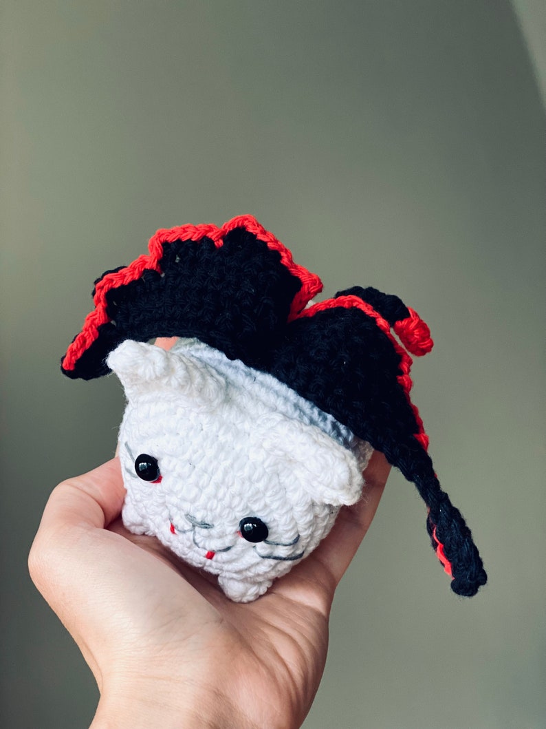 Puede incluir: Un gato blanco de crochet con una capa de crochet negra y roja. El gato tiene ojos negros y una boca roja.