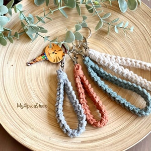 May include: Four crocheted keychains in gray, blue, brown, and white. The keychains are hanging from a wooden plate with the text "MyAlpacaStudio" on it.