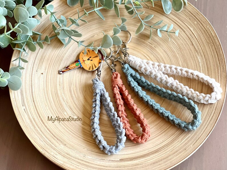 May include: Four crocheted keychains in gray, blue, brown, and white. The keychains are hanging from a wooden plate with the text "MyAlpacaStudio" on it.