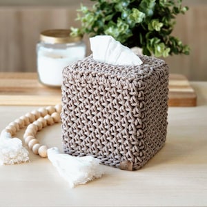 May include: A square tissue box cover in a neutral beige color, featuring a woven texture. The top opening reveals white tissues. Decorative wooden beads with white tassels are in the foreground. A candle in a glass jar is in the background.