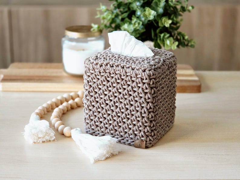 May include: A square tissue box cover in a neutral beige color, featuring a woven texture. The top opening reveals white tissues. Decorative wooden beads with white tassels are in the foreground. A candle in a glass jar is in the background.